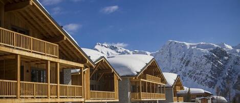 Una cabaña de madera con un balcón, frente a un paisaje montañoso cubierto de nieve.