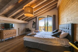 Quarto com uma cama grande, televisão sobre uma cômoda de madeira, e varanda com vista para árvores.