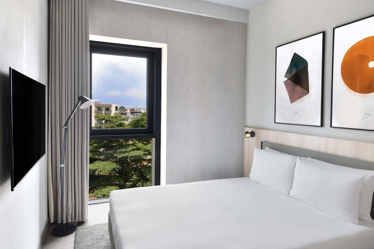 A modern hotel room with a large bed, a flat-screen TV, a window with curtains, and two framed artworks on the wall.