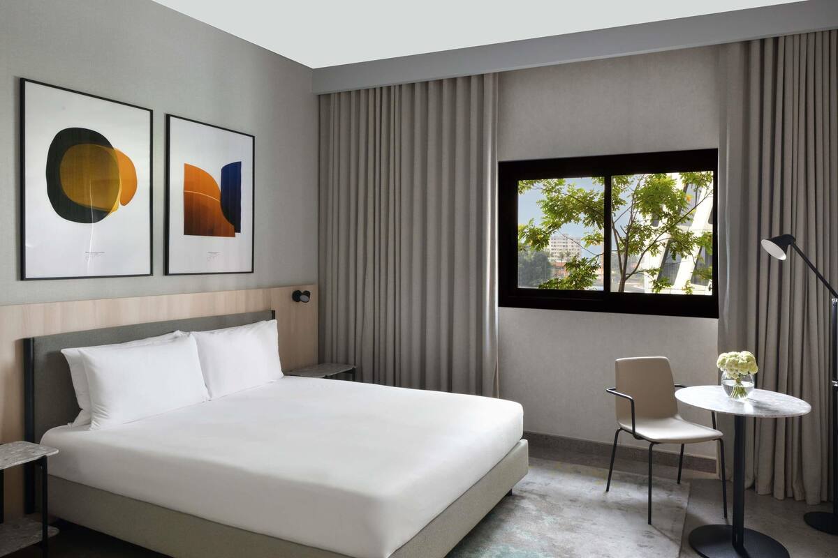 Ett modernt hotellrum med en stor säng, ett litet bord, en stol och ett fönster med utsikt över byggnader och träd.