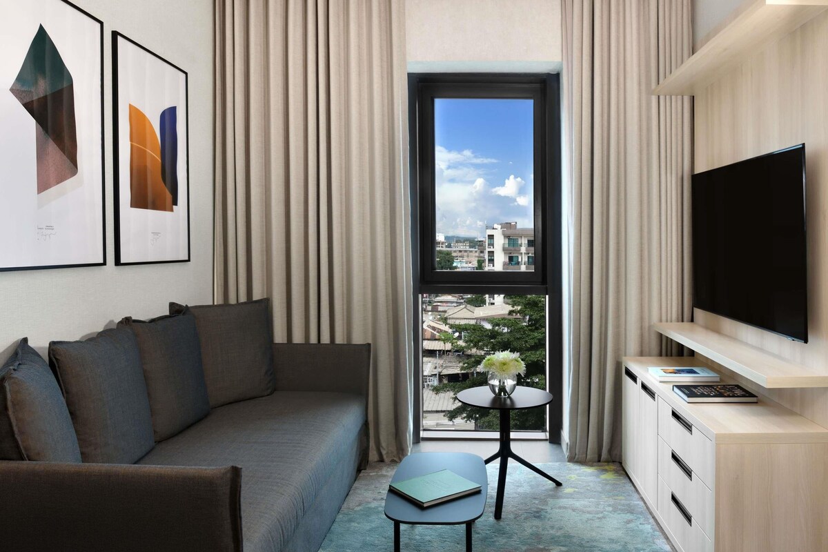 Een moderne hotelkamer met een bank, een klein tafeltje met een vaas, een flatscreen televisie en een raam met uitzicht op de stad.