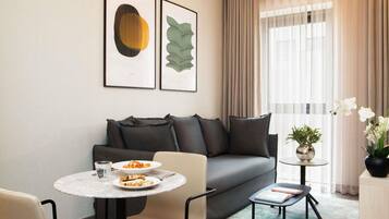 Une chambre d’hôtel moderne avec un canapé gris, une table de salle à manger ronde, une petite table d’appoint et une fenêtre avec des rideaux.