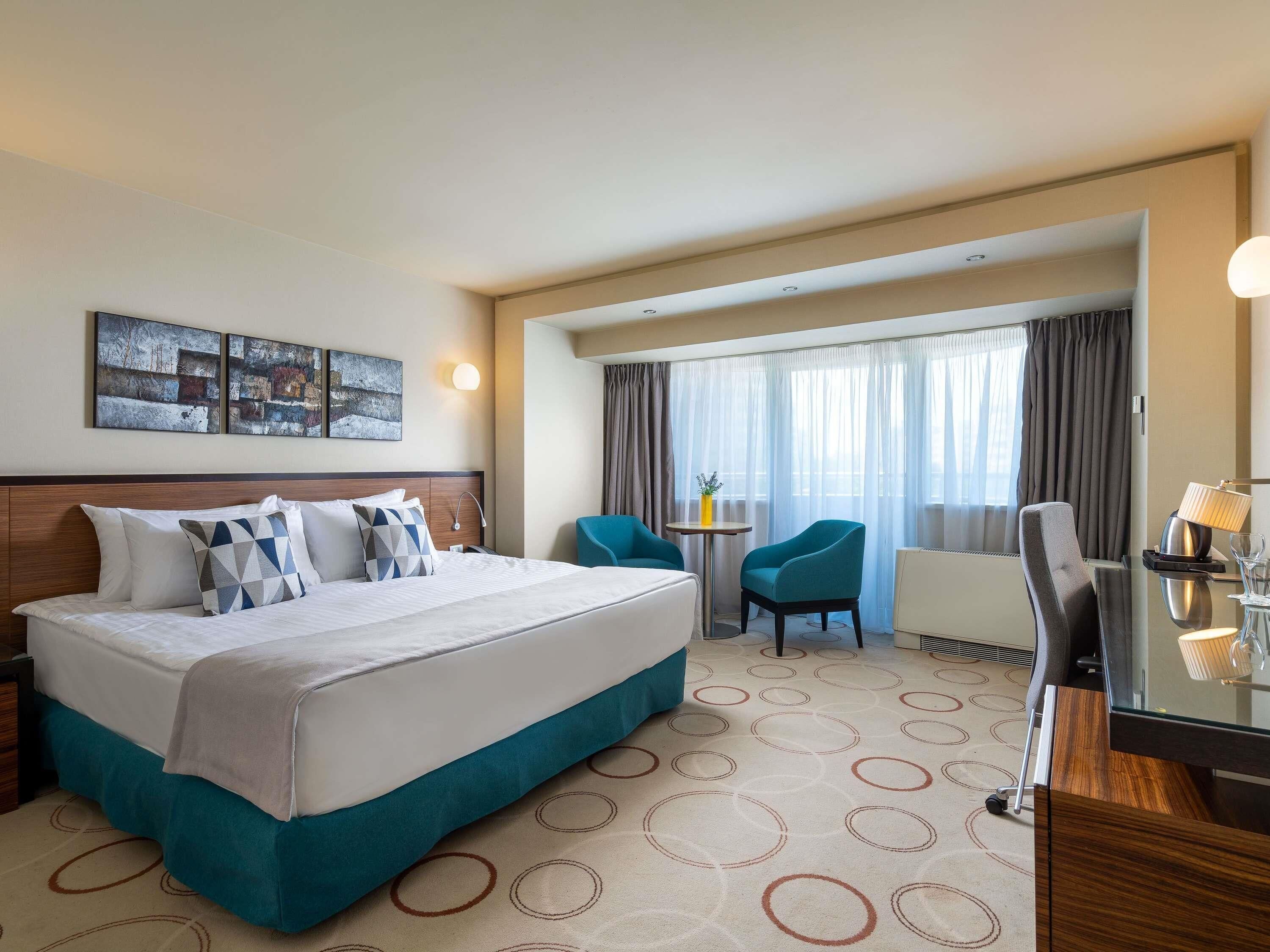 A hotel room with a bed, a desk, a chair, a small table, and a window with curtains.