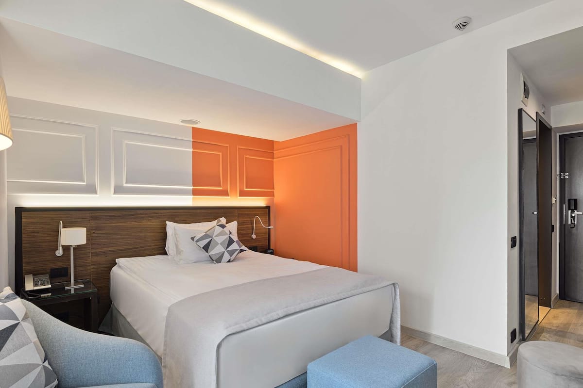 Une chambre d’hôtel moderne, équipée d’un lit, de deux fauteuils, d’une table de chevet et d’une porte donnant sur une autre pièce.