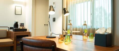A hotel room with a wooden desk, a leather chair, a bedside table with a lamp, and a window with blinds.