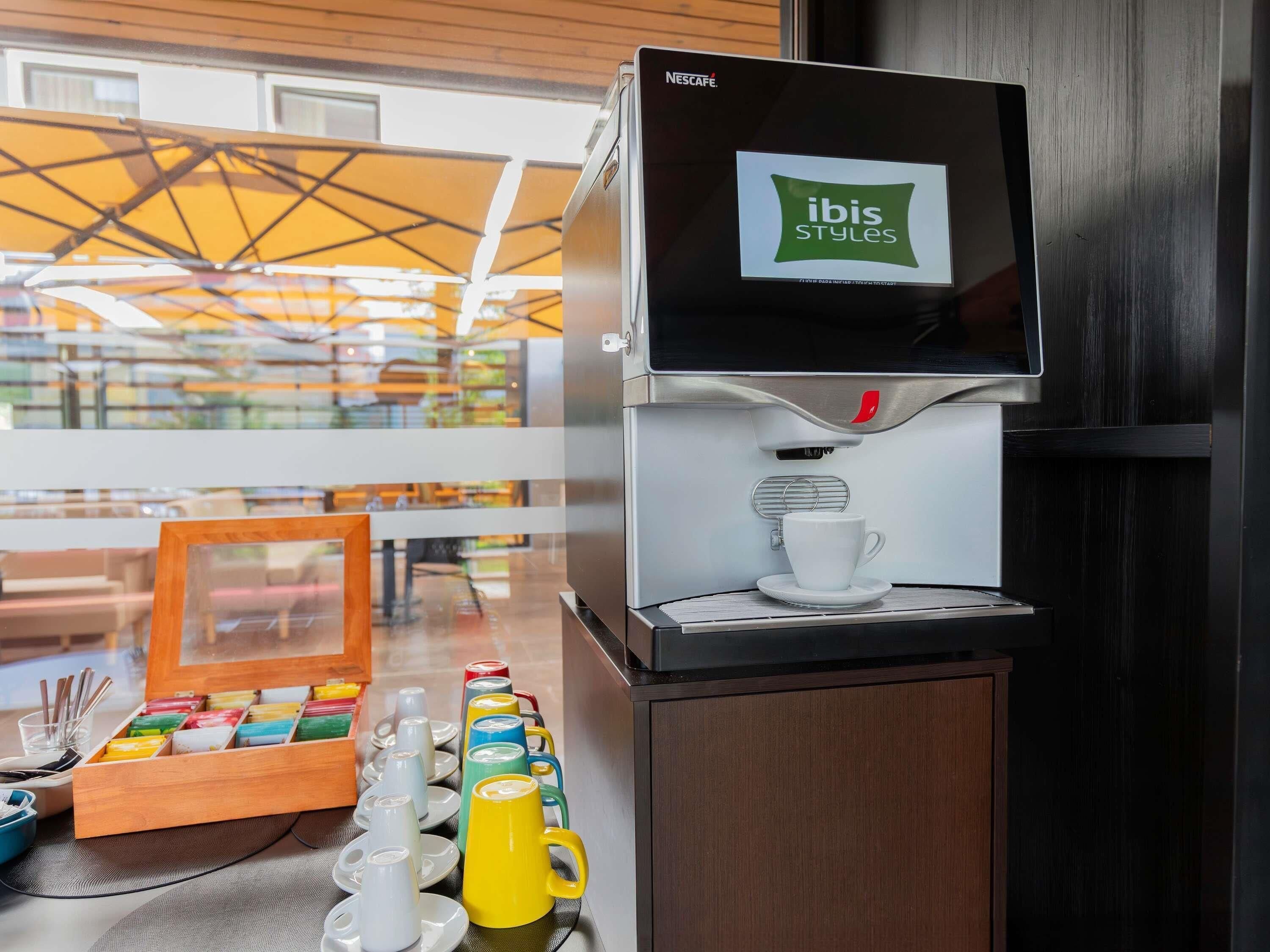 Una máquina de café Nescafé con un rótulo de 'Ibis Styles', una taza de café y una variedad de tazas y platillos de colores sobre una mesa.