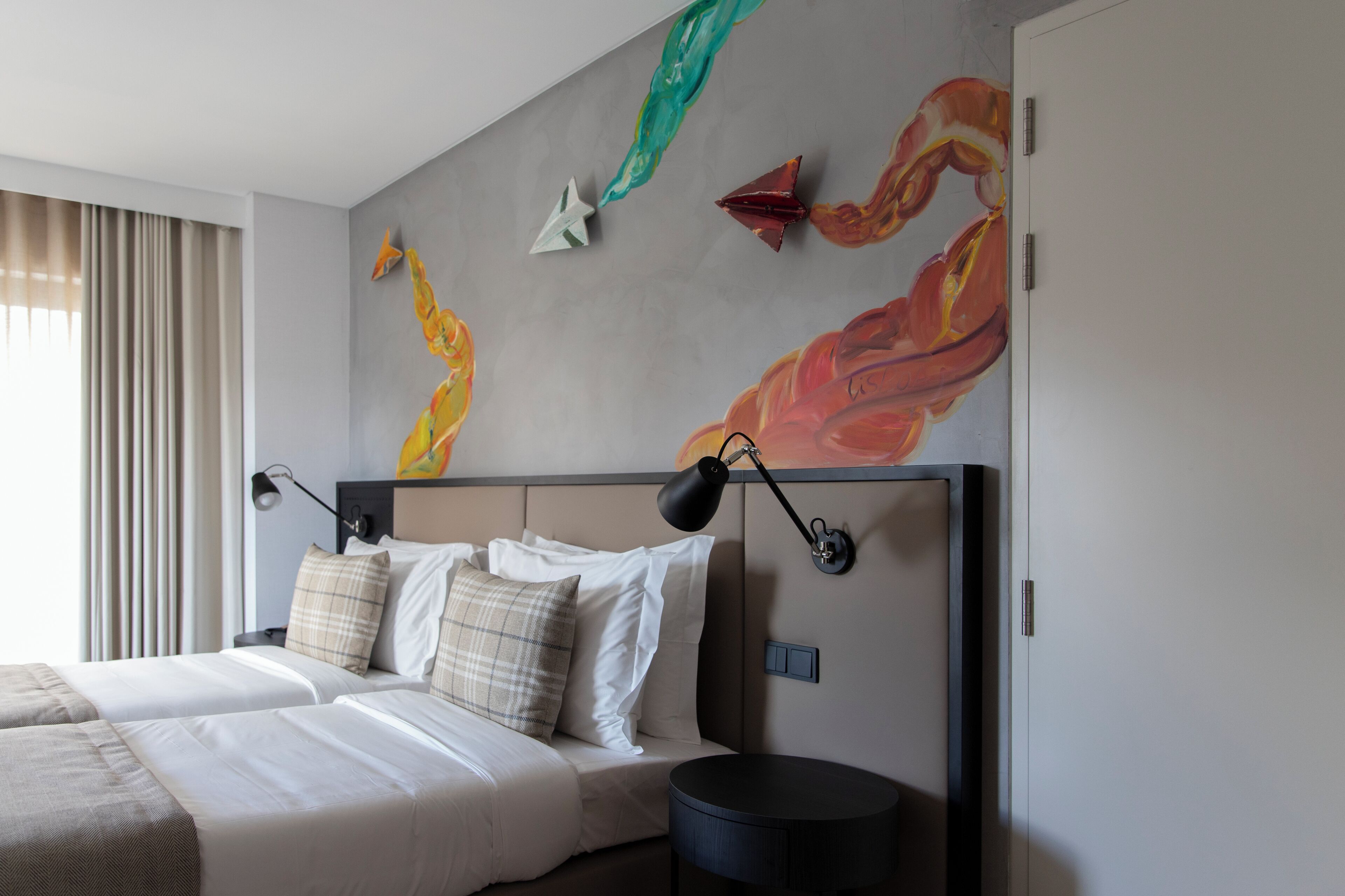 Una habitación de hotel con una cama grande, dos mesitas de noche, una lámpara de pared y un cuadro con formas abstractas.