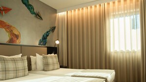 A hotel room with two beds, a headboard, a nightstand, a lamp, and a large window with curtains. - ibis Styles Lisboa Aeroporto (Loures)