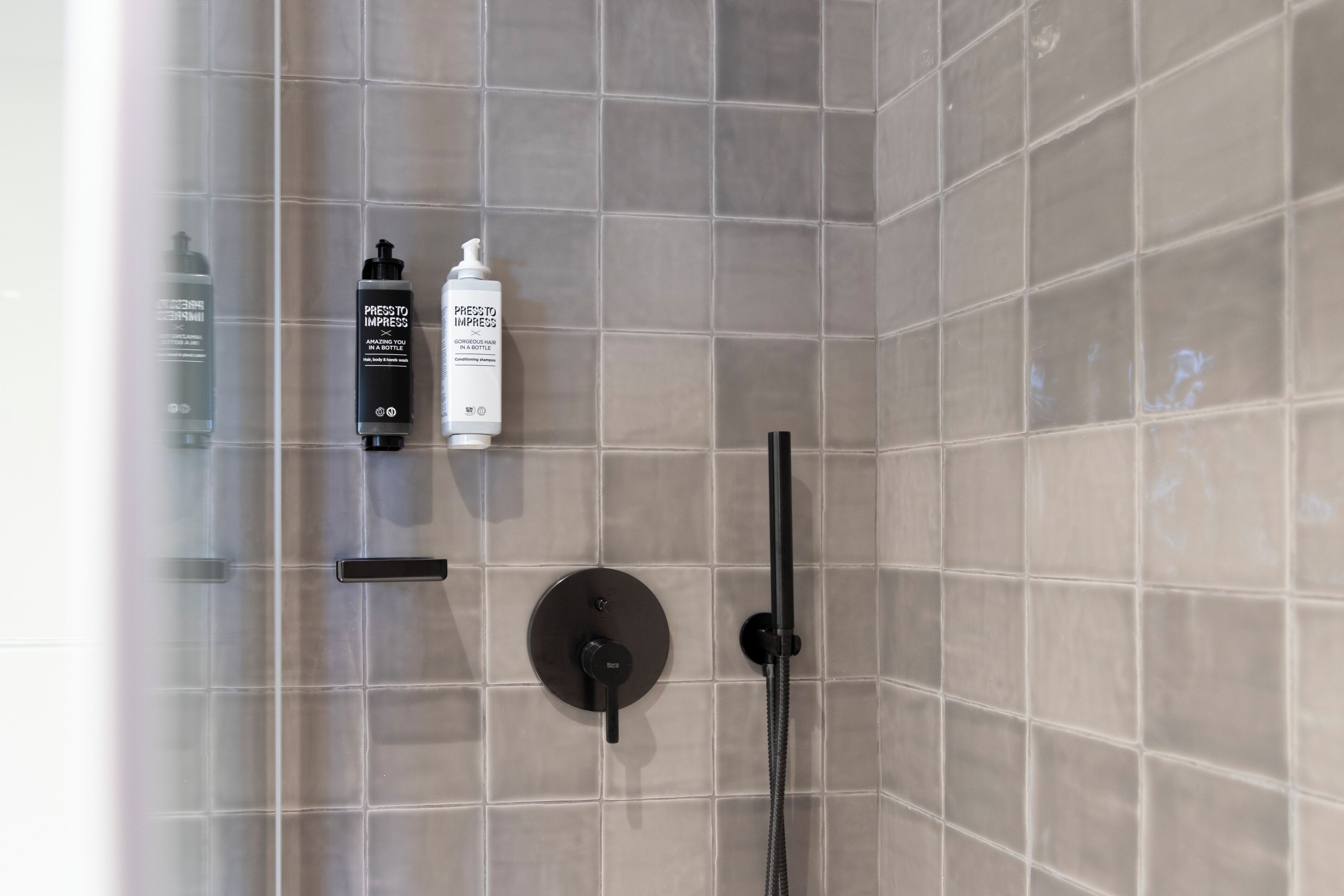 Zona de ducha con cabezal de ducha de mano, panel de control en la pared y dos botellas de champú y acondicionador.