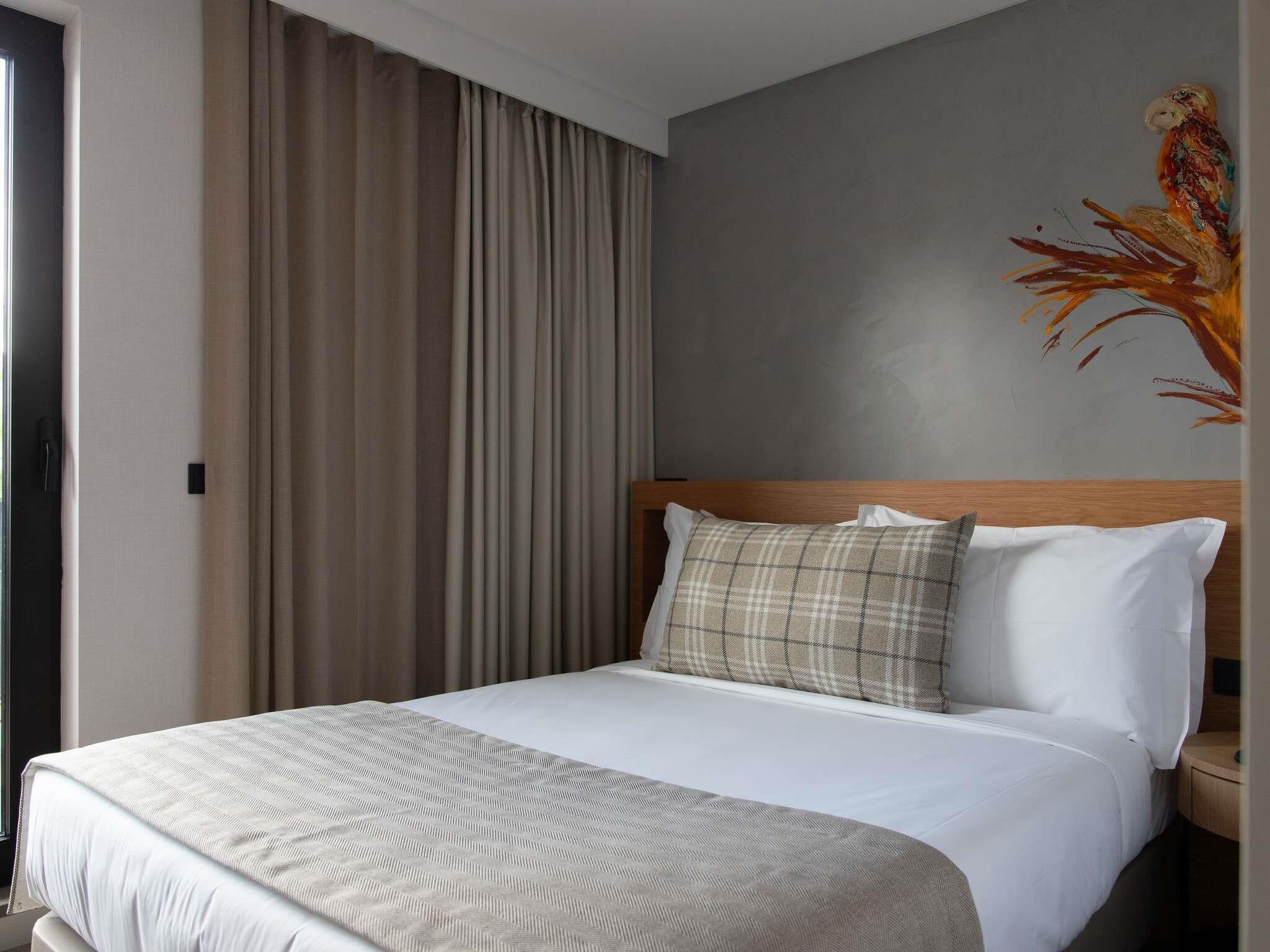 A hotel room with a bed, curtains, a headboard, and a painting on the wall.