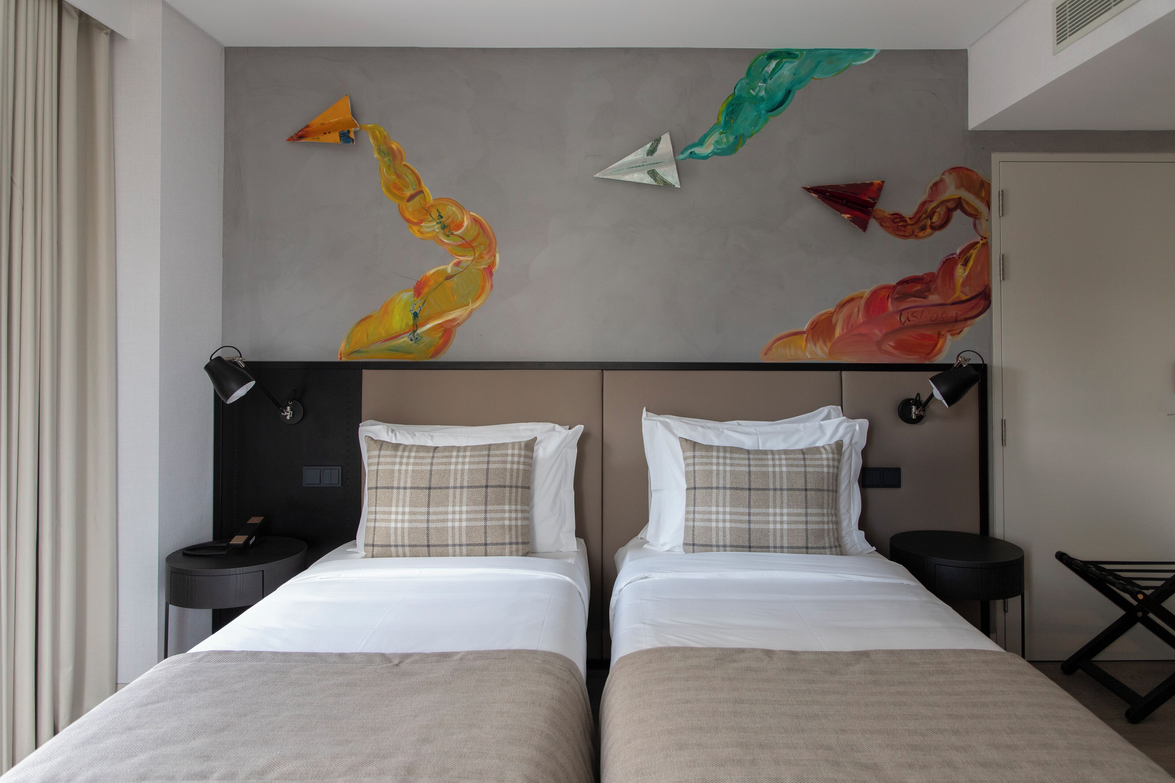 Quarto de hotel com duas camas, uma grande pintura abstrata na parede e uma mesa redonda pequena com uma luminária.