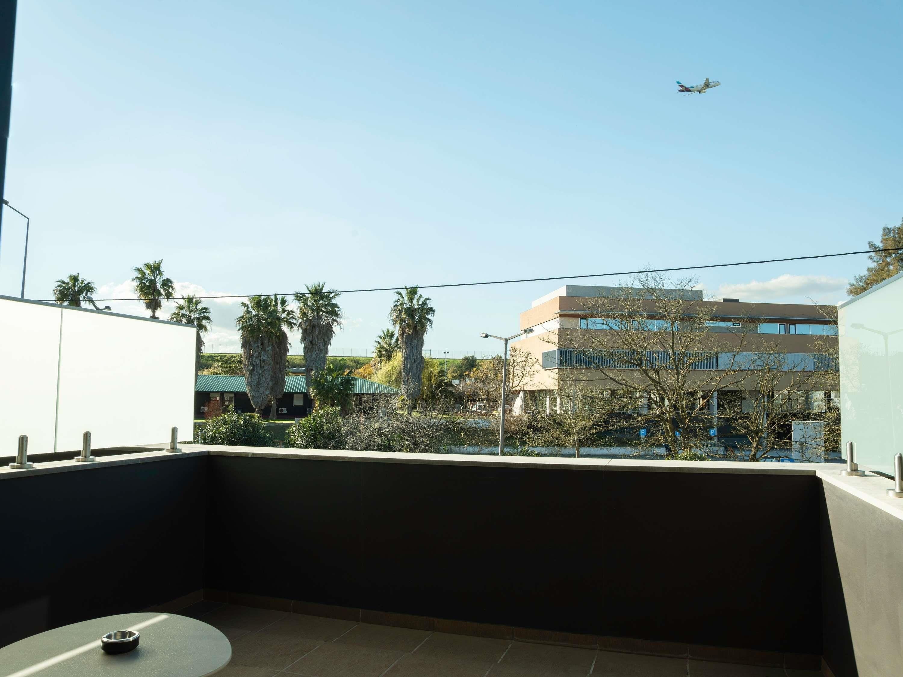 A balcony with a view of a building, palm trees, and an airplane in the sky.