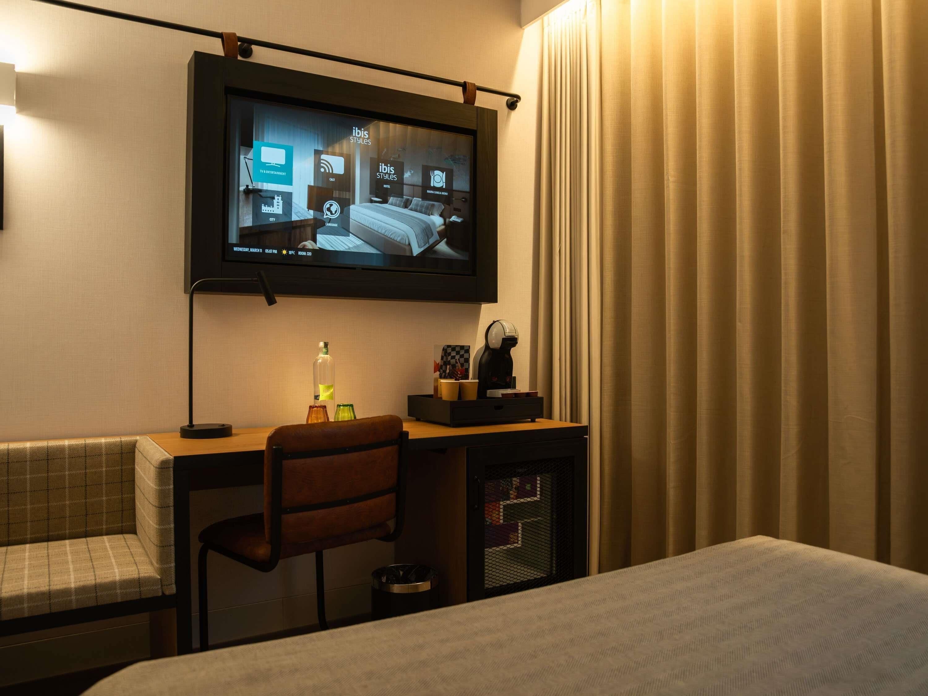 Una habitación de hotel con cama, escritorio con silla, televisión montada en la pared y un minibar.