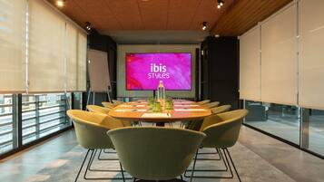 Un salón de conferencias con una pantalla grande mostrando 'ibis Styles', una mesa redonda con sillas y vistas al exterior a través de amplios ventanales.