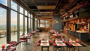 Un restaurante con techos altos, amplios ventanales y vistas al paisaje. Las mesas están dispuestas con servilletas rojas y vasos.