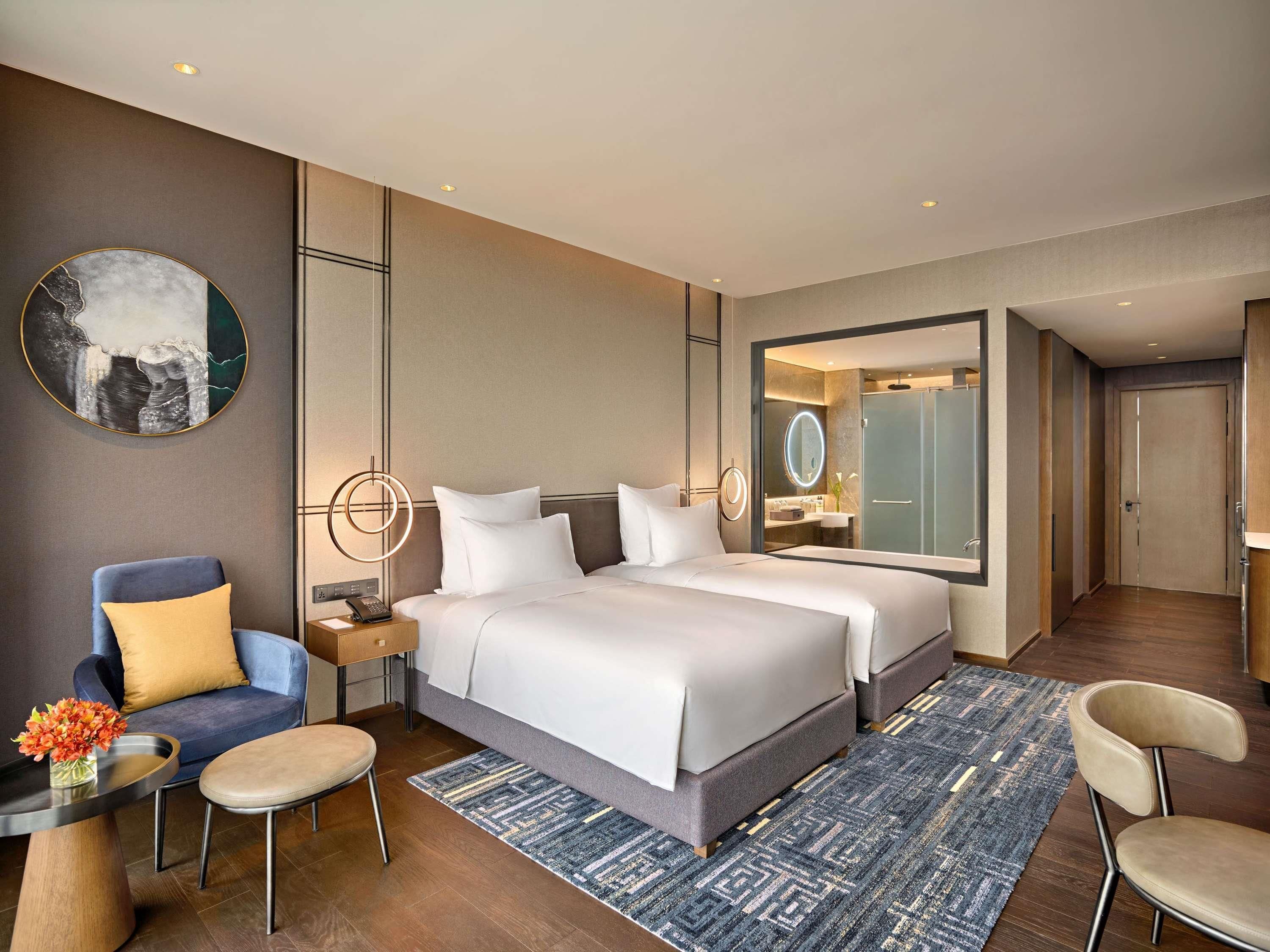 A modern hotel room with a large bed, a desk with a phone, a chair, a round mirror, and a view of the bathroom.