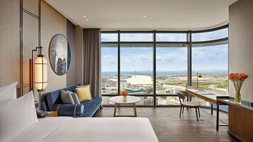 A modern hotel room with a large bed, a sofa, a dining table, and a view of a cityscape through large windows.