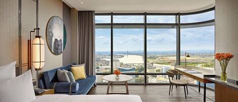 A modern hotel room with a large bed, a sofa, a dining table, and a view of a cityscape through large windows.