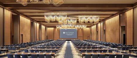 Un amplio salón de conferencias con filas de mesas y sillas, un escenario con una pantalla de presentación y modernas lámparas de araña.