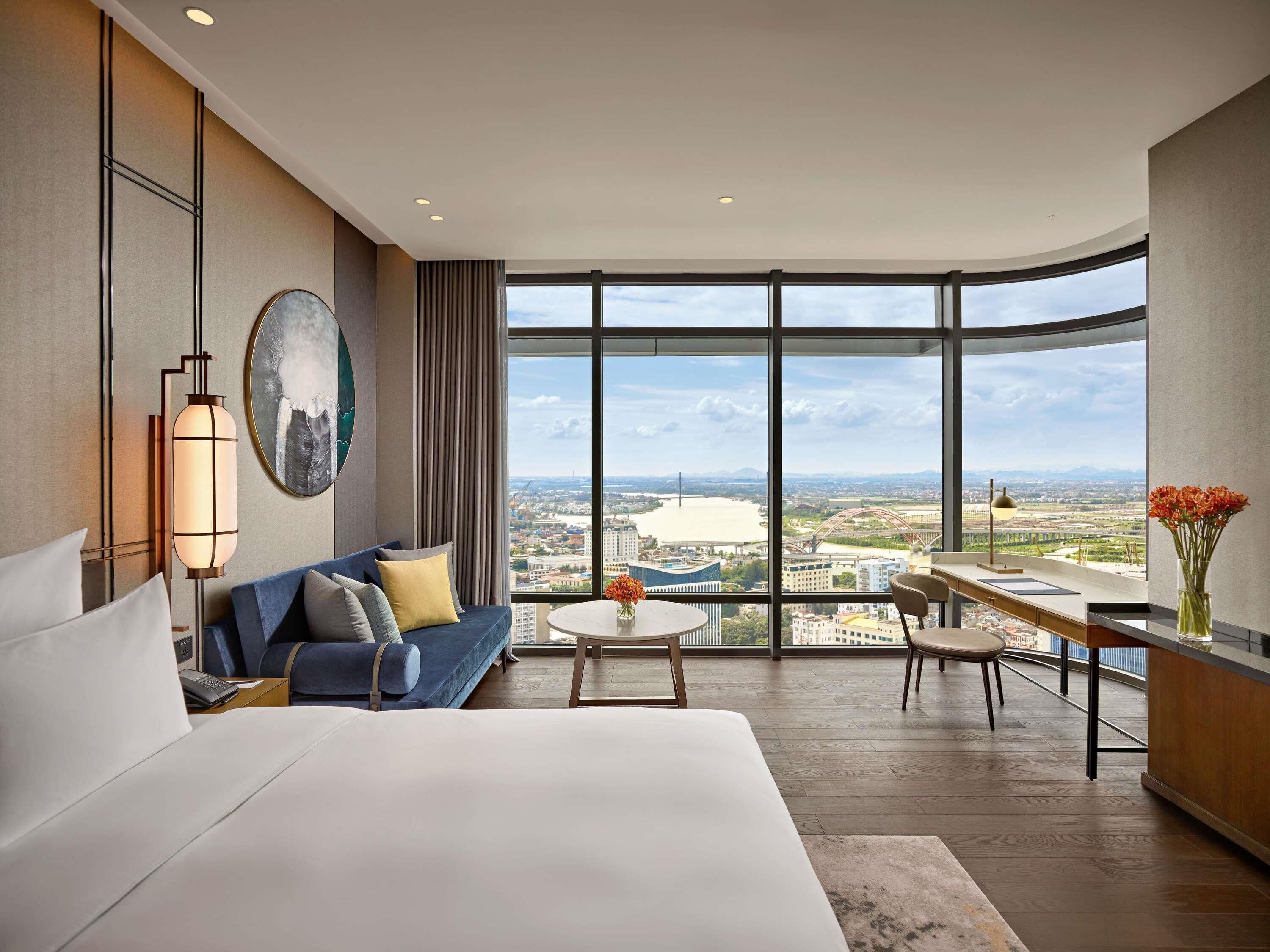 Una habitación de hotel moderna con una cama grande, un sofá, una mesa de comedor y vistas a un paisaje urbano a través de amplios ventanales.