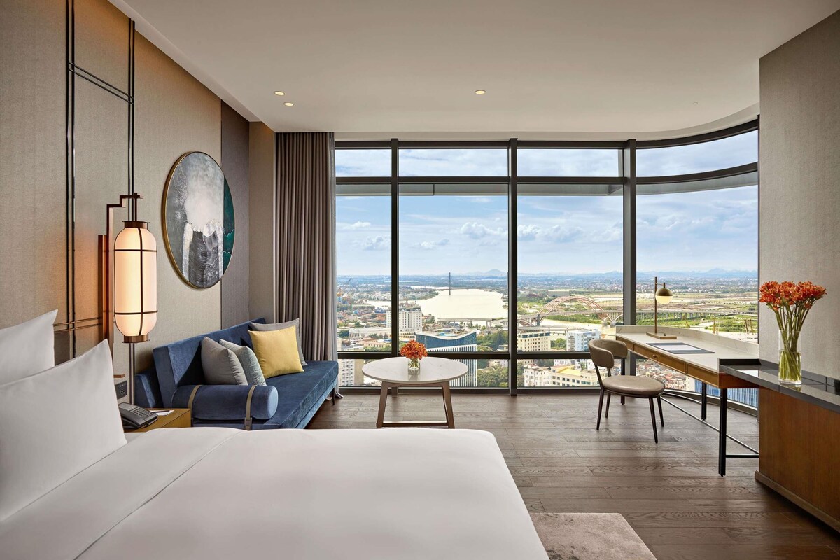 Una habitación de hotel moderna con una cama grande, un sofá, una mesa de comedor y vistas a un paisaje urbano a través de amplios ventanales.