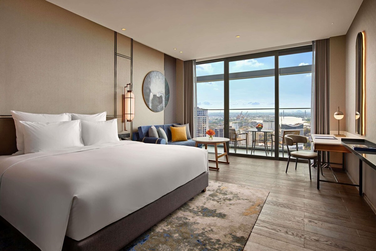 Habitación de hotel con una cama grande, un escritorio, una silla, una mesita con un jarrón y vistas a la ciudad.