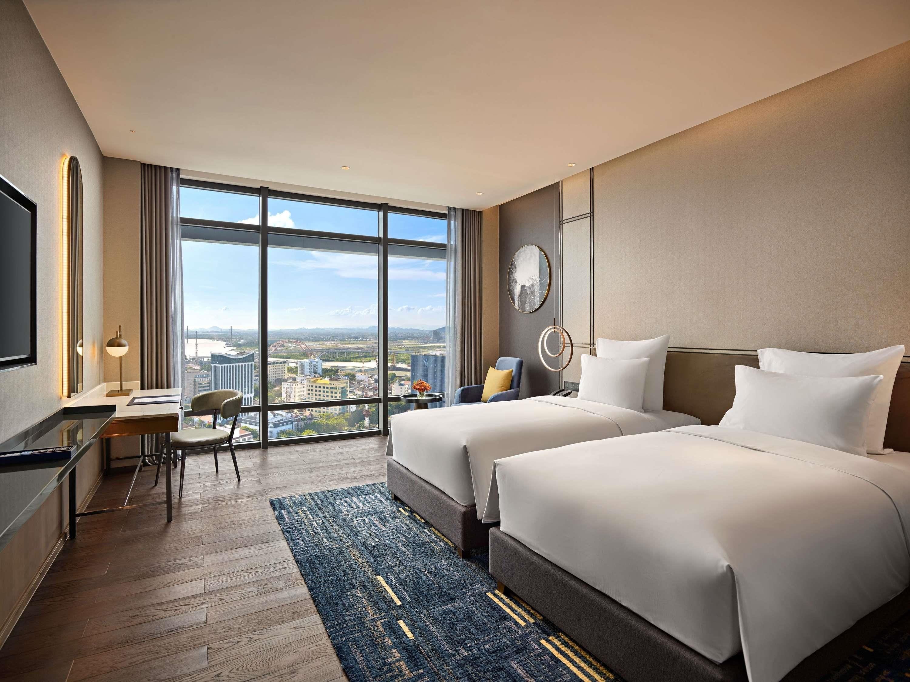 Habitación de hotel con dos camas, un escritorio, una silla y un amplio ventanal con vista a la ciudad.