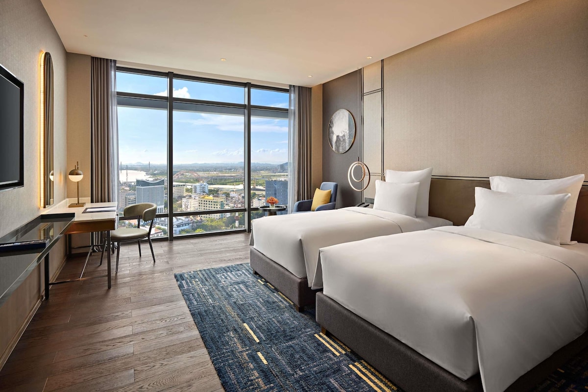A hotel room with two beds, a desk, a chair, and a large window offering a city view.