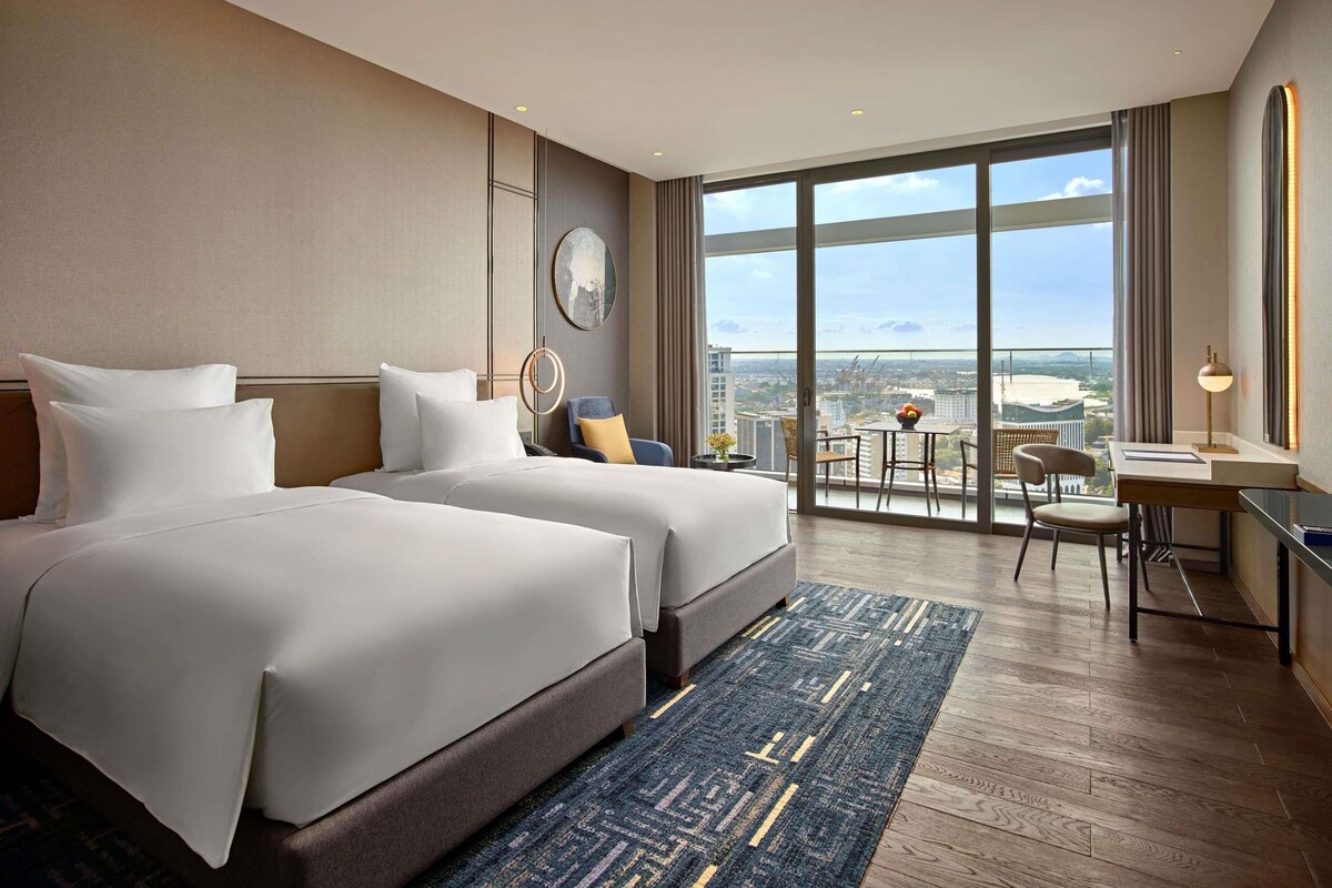 Habitación de hotel con dos camas, un escritorio y un ventanal con vistas a la ciudad.