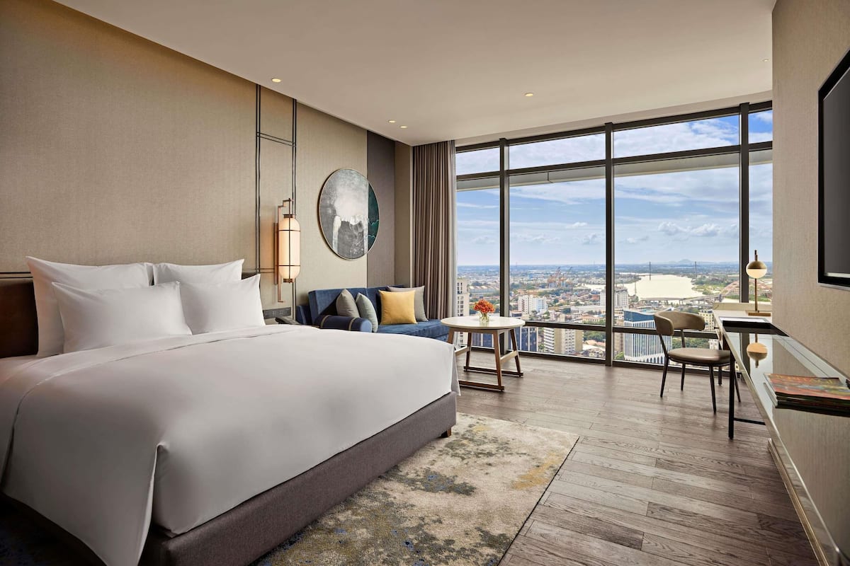 A hotel room with a large bed, a desk, a chair, a round table, and a large window with a city view.