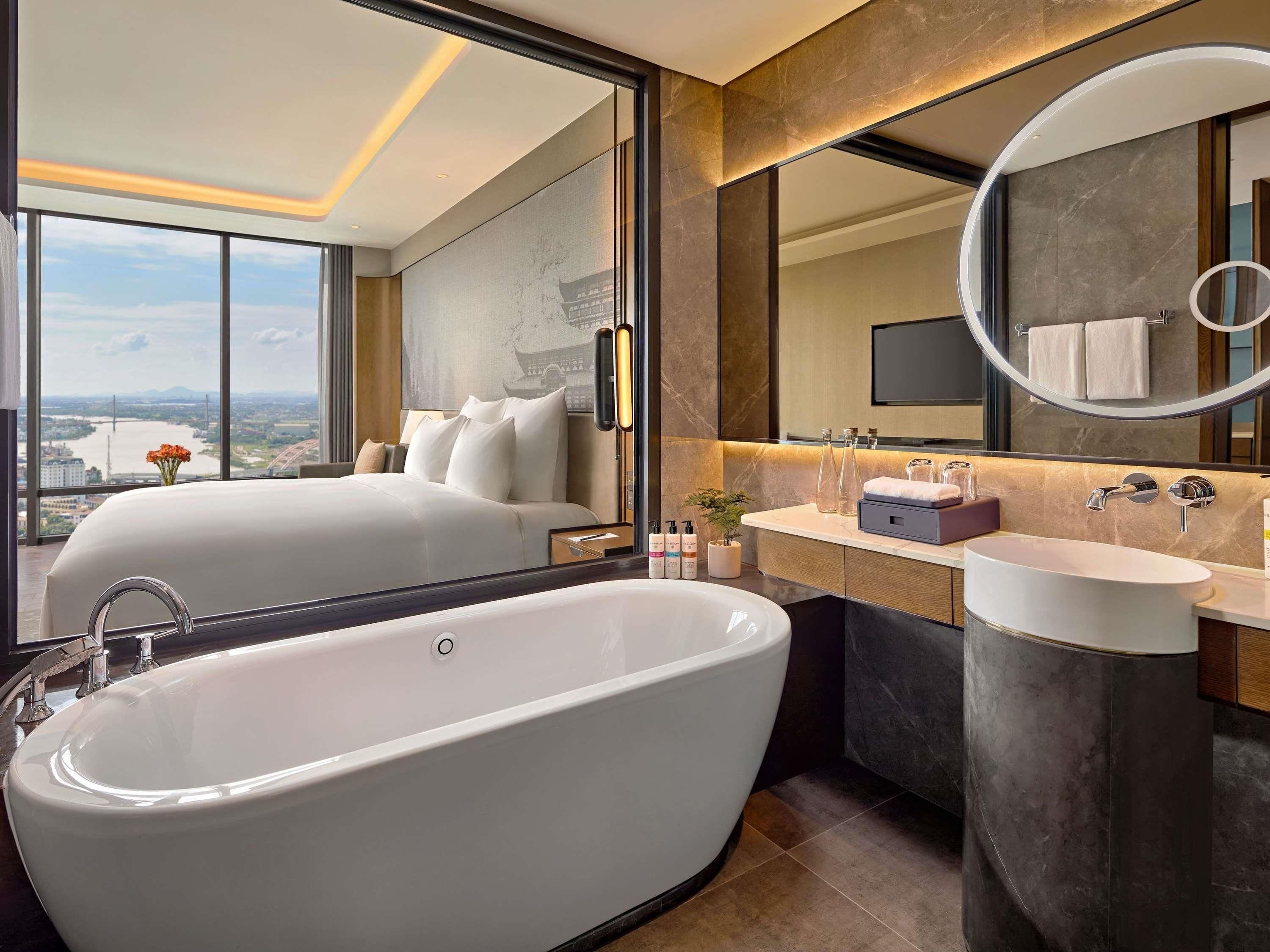 A modern hotel room with a large bathtub, a vanity with a mirror, and a bed with a city view.