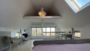 Une chambre spacieuse située dans les combles, avec un grand lit, un lustre et une vue sur l’extérieur grâce à de grandes fenêtres.