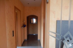 Un couloir aux murs en bois, une porte sur la gauche, une petite fenêtre au fond et un sol carrelé.