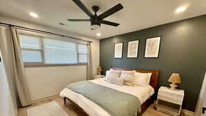 Et soveværelse med seng, ventilator, natbord, lampe og et vindue med persienner.