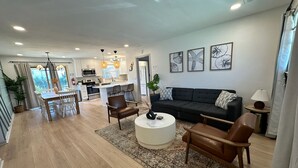 En rummelig opholdsstue med sofa, stole, sofabord og spisebord. I baggrunden er der et køkken med moderne hårde hvidevarer og et spiseområde med et stort vindue.