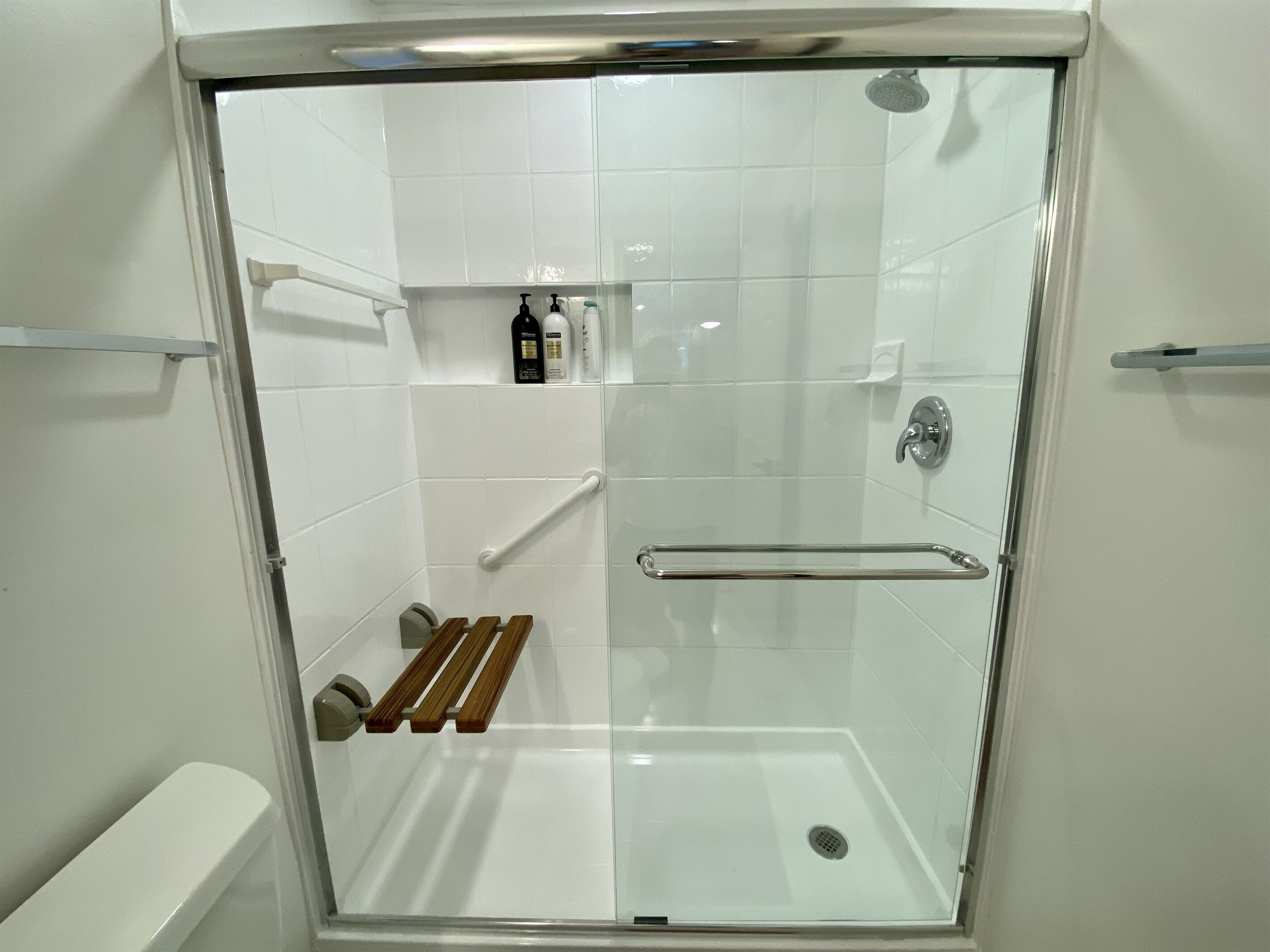 Une douche fermée par des parois en verre, avec un mur recouvert de carreaux blancs, une pomme de douche à main, un distributeur de savon à main et un tapis de bain en bois.