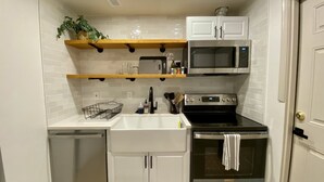 A compact kitchen with white cabinets, a microwave, an oven, and a sink. There are shelves above the counter holding glassware and a plant.
