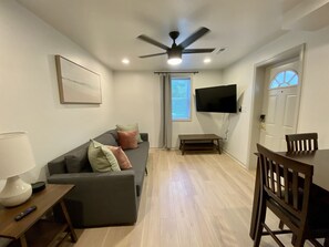 A living room with a grey sofa, a wooden coffee table, a TV mounted on the wall, and a ceiling fan.