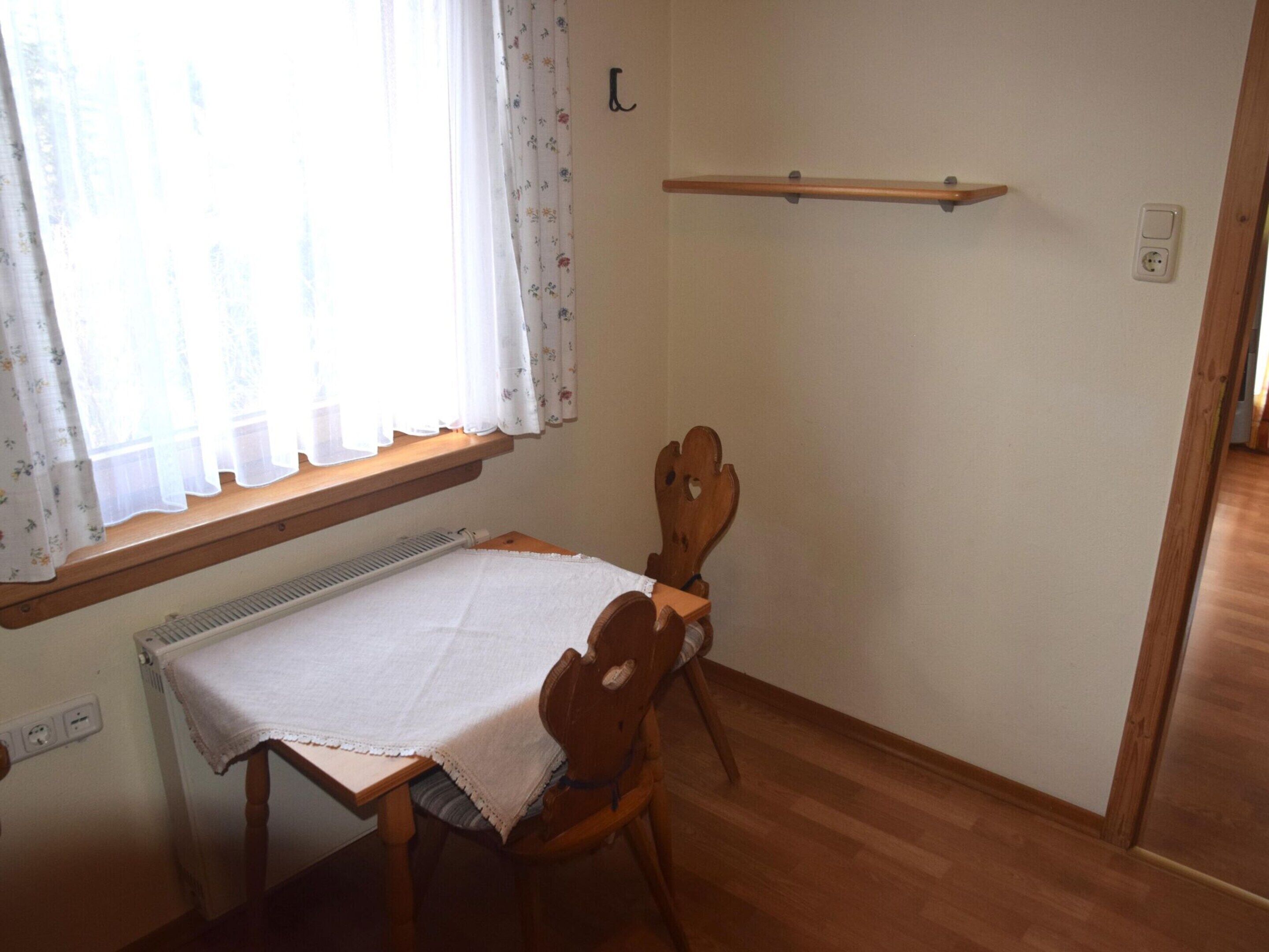 Una habitación pequeña con una mesa y sillas de madera, un radiador debajo de la ventana y un estante de madera sobre la mesa.