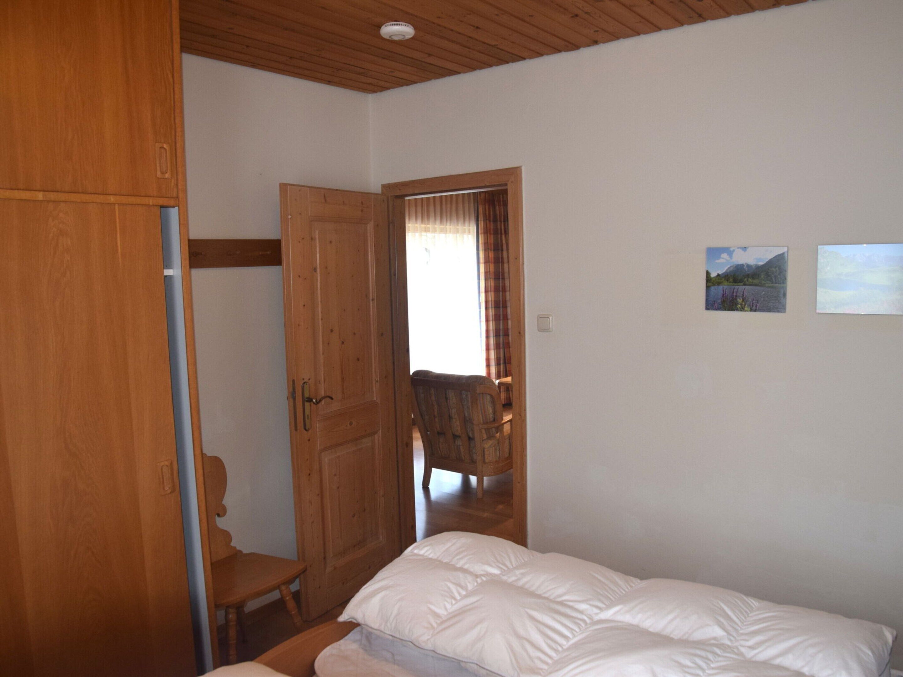 A bedroom with a wooden wardrobe, a bed with white bedding, a wooden chair, and two framed pictures on the wall.