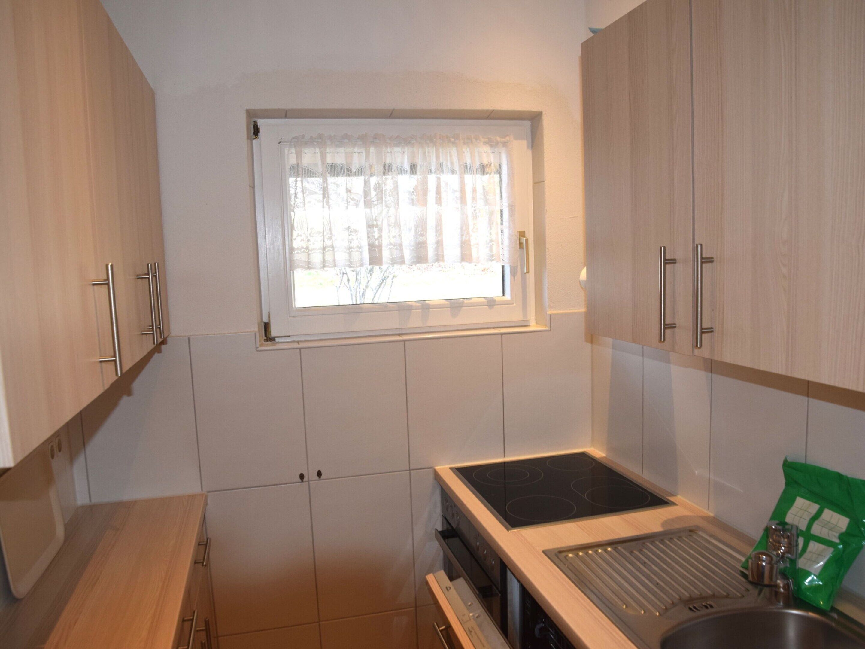 A compact kitchen with wooden cabinets, a window with a sheer curtain, and a stainless steel sink.