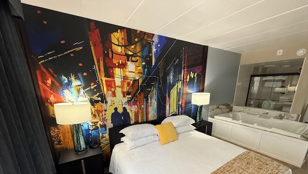 A hotel room with a large bed, two bedside lamps, a modern bathroom, and a vibrant abstract painting on the wall.