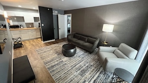 A modern living room with a grey sofa, a white armchair, a coffee table, and a patterned rug. There is a kitchen in the background with white cabinets and a black door. - Lighthouse Inn & Suites (Hannibal)