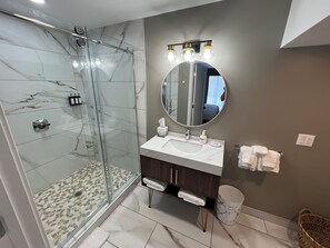 A modern bathroom with a glass shower, marble tiles, a round mirror, and a vanity with a sink and towel rack.