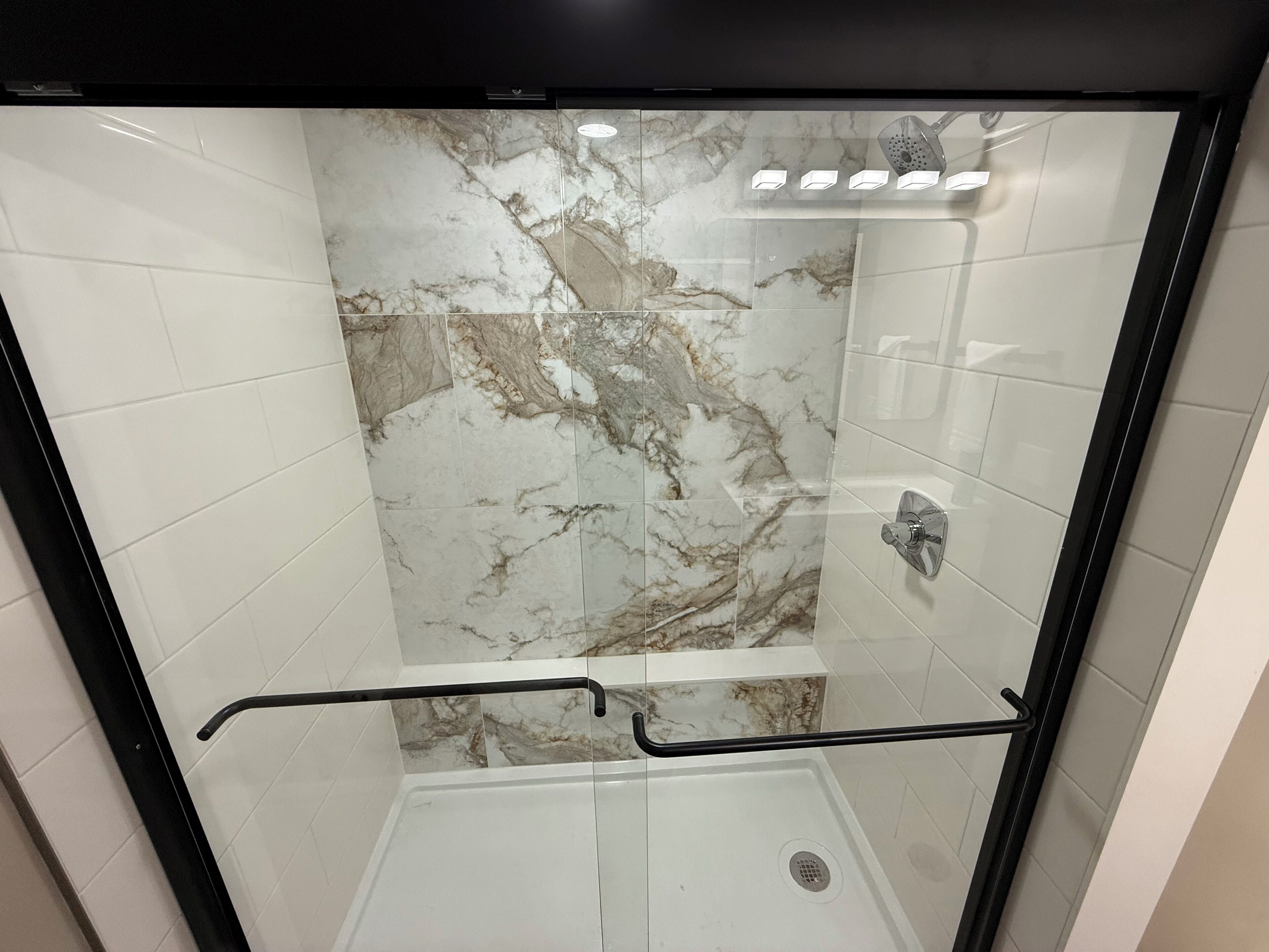 A glass-enclosed shower with a marble wall and a black handrail.