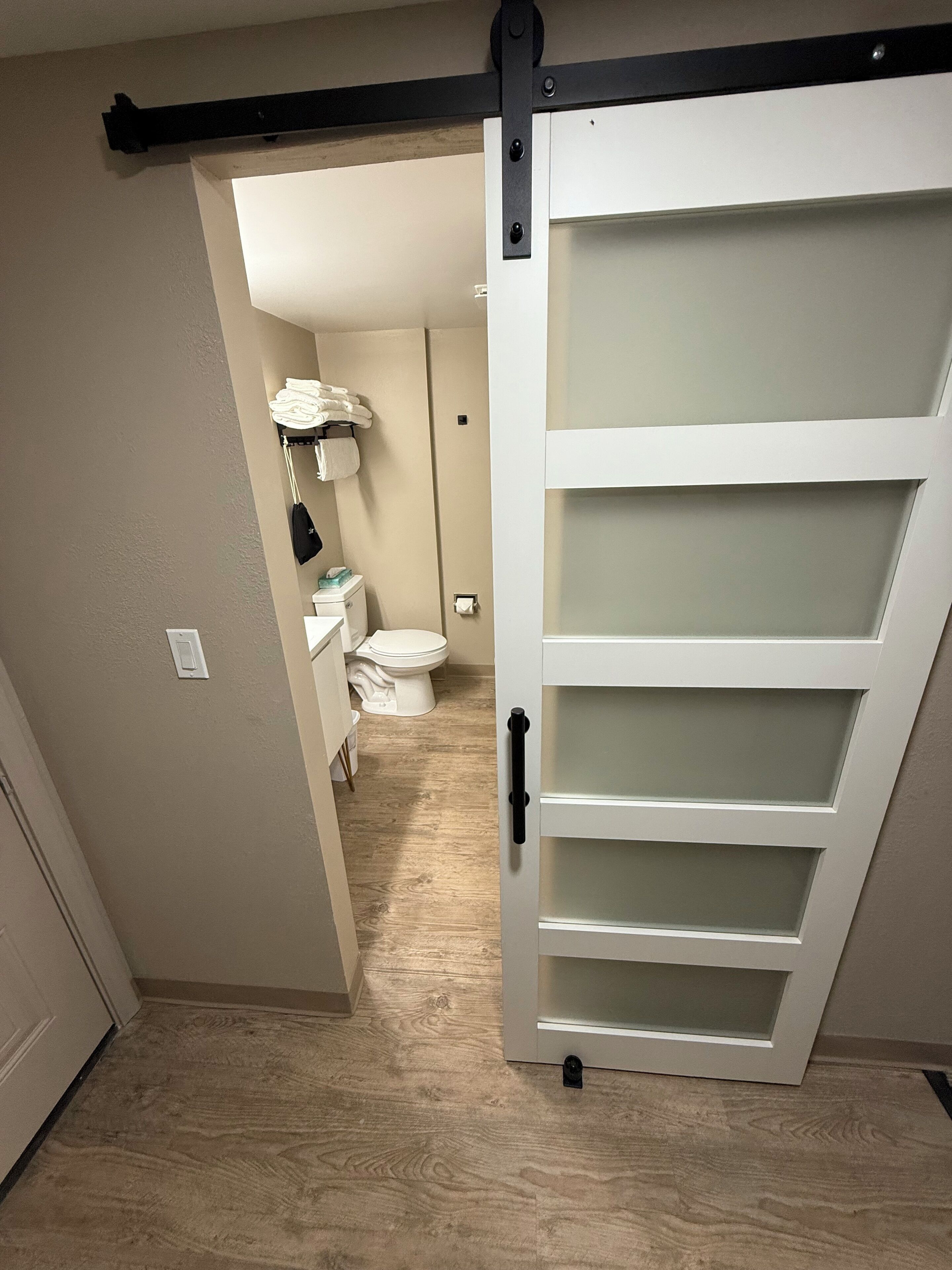Un couloir menant à une salle de bain équipée de toilettes, d’un porte-serviettes et d’une armoire blanche.