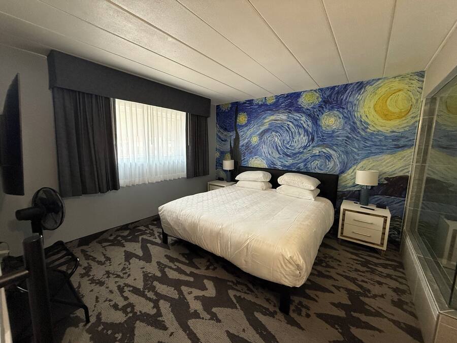 A hotel room with a bed, a nightstand, a lamp, a fan, a window with curtains, and a wall with a famous painting.