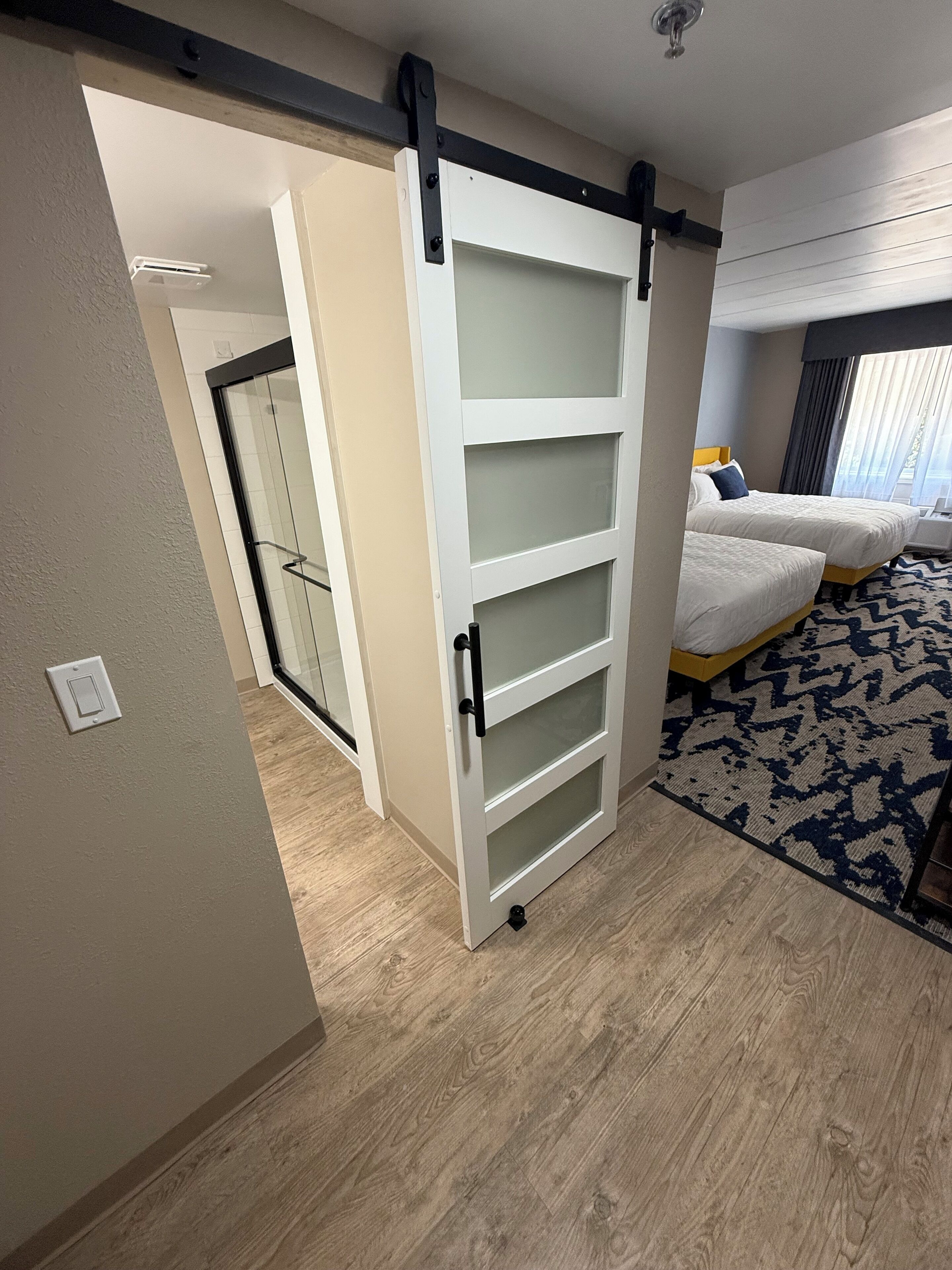 Habitación de hotel con una puerta corredera que da a un baño, una cama con cabecera y un tapete estampado.