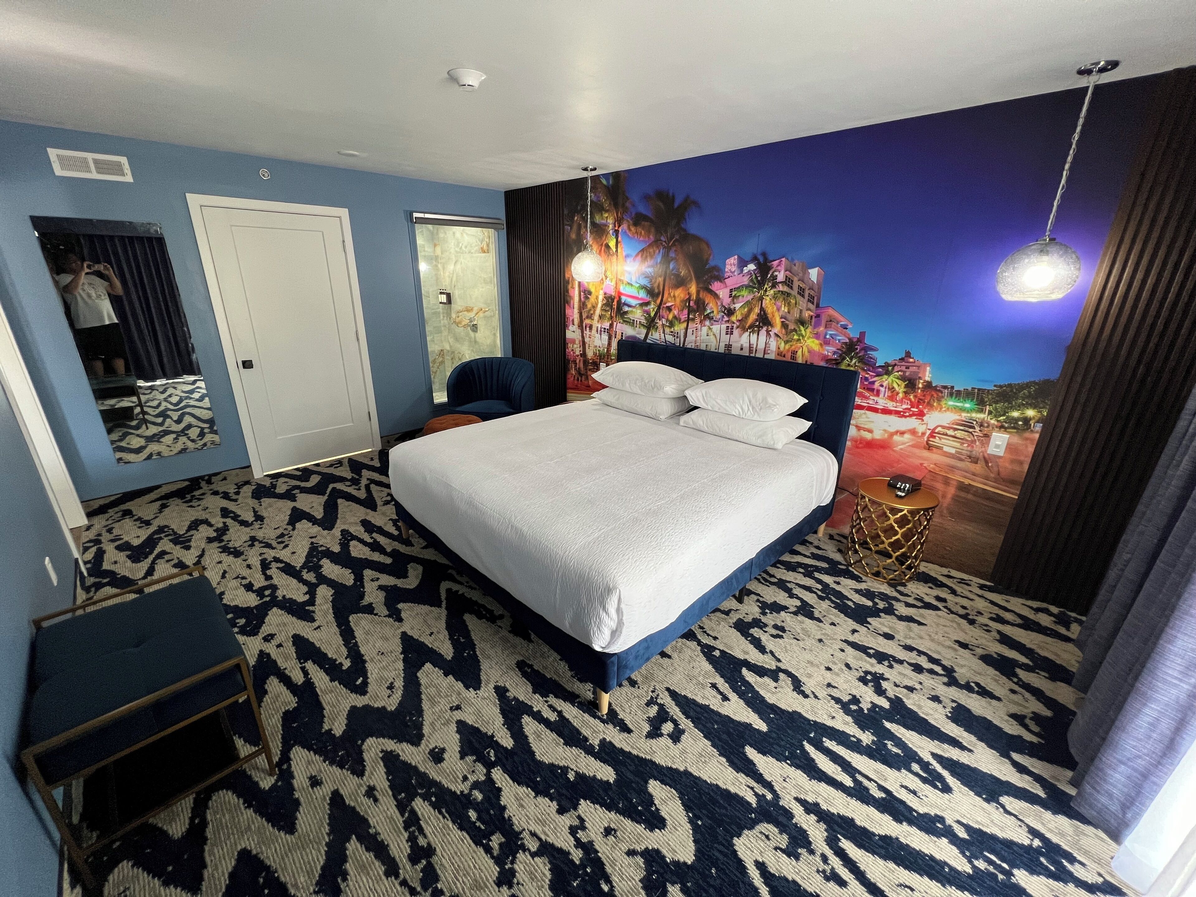 Une chambre d’hôtel avec un lit, une chaise, une petite table, un miroir et une fresque murale.