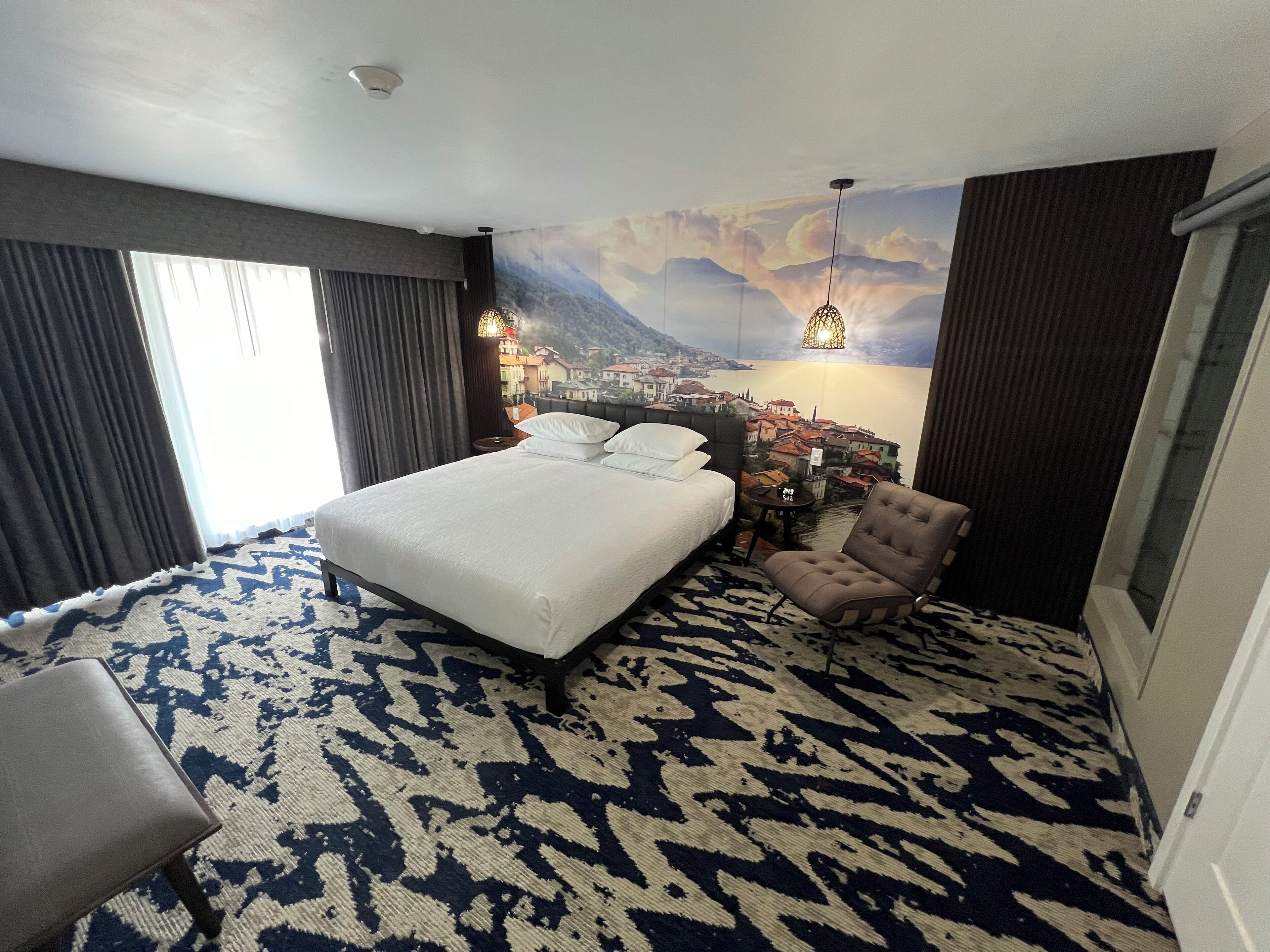 A hotel room with a large bed, a chair, a patterned carpet, a mural of a town, and a window with curtains.
